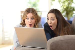 Front view portrait of two surprised friends watching on line content in a laptop lying on a couch at home