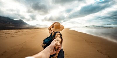 Happy and excited girl taking her boyfriend's hand while walking enjoying summer sunset at the beach - Traveler couple leads on vacation between the mountain - Follow me concept and traveling together